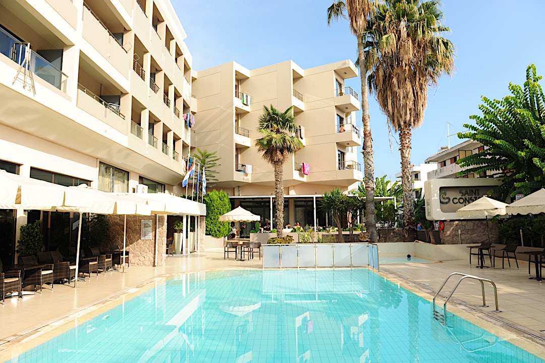 Pool med stenlagd däckyta och beige hotellbyggnad med balkonger i bakgrunden.