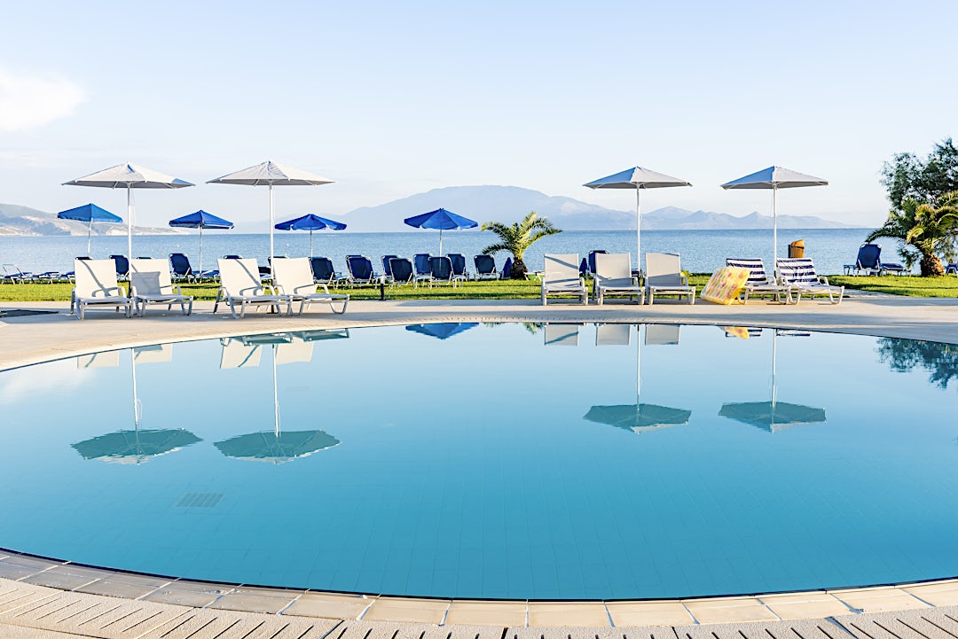 Cirkulär blå pool med vita parasoller, palmer, berg och havet i bakgrunden.