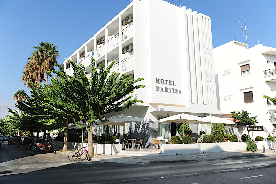 Exteriör vy av vit hotellbyggnad med palmer och vita parasoller i förgrunden.