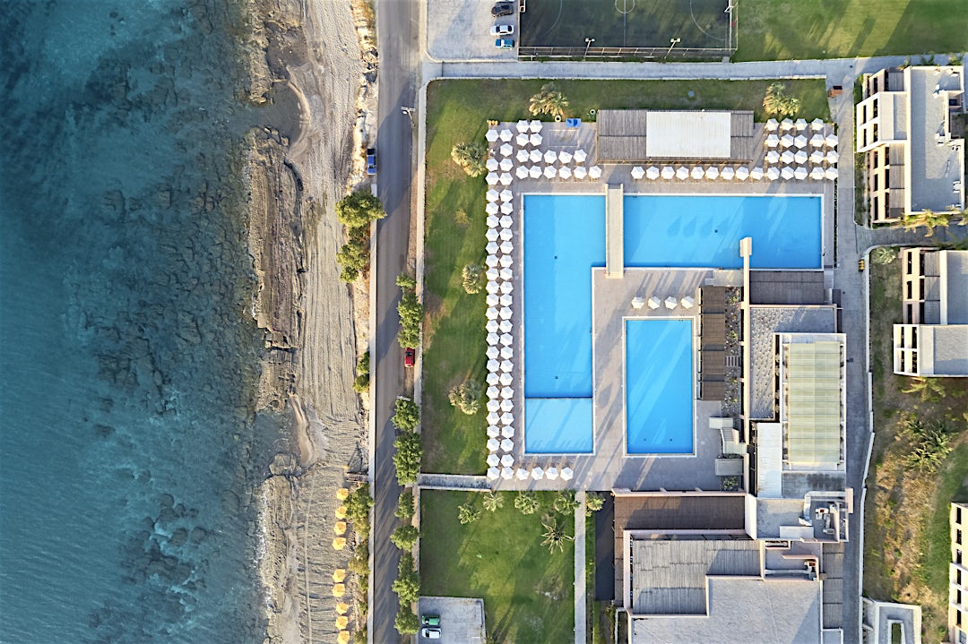 Flygvy över kustresort med turkosa pooler intill havet, vita byggnader och sandstrand.