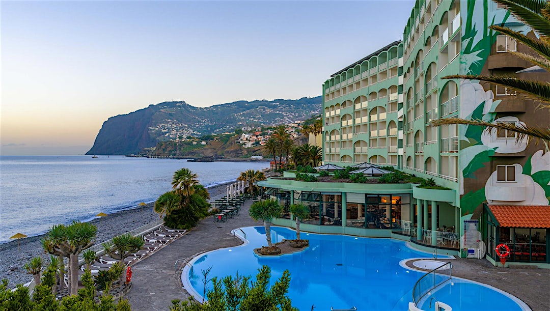Hotellbyggnad med pool framför, utsikt över hav och berg i bakgrunden.