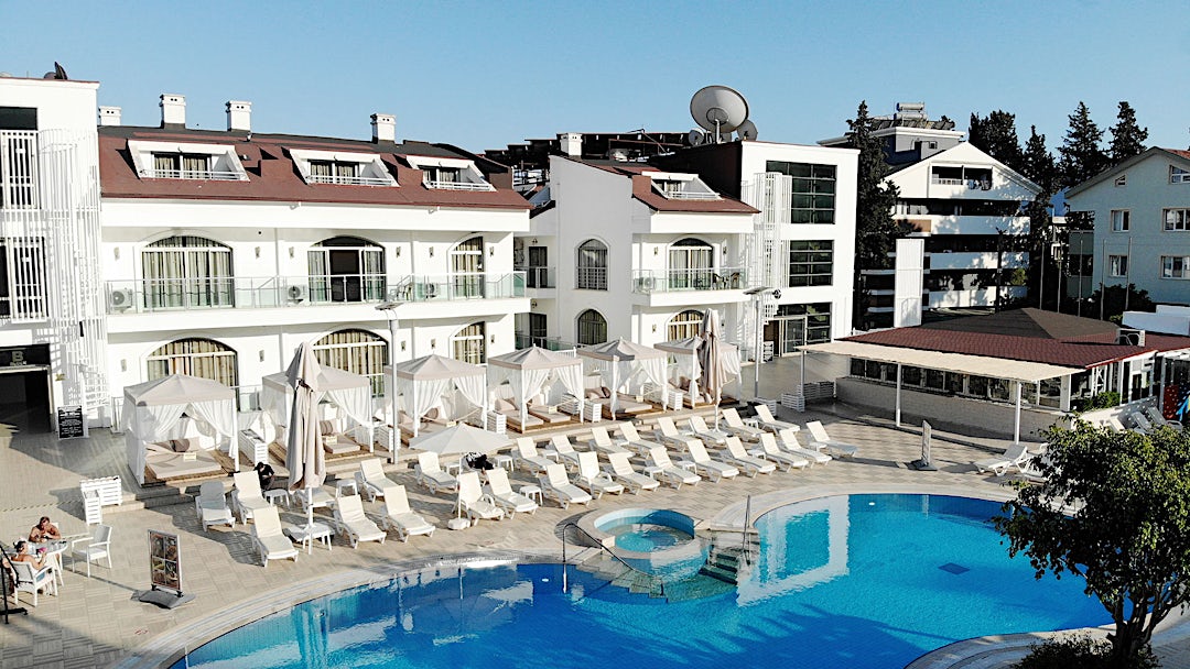 Ljus hotellfasad i bakgrunden med pool och solstolar i förgrunden.